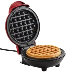 Máquina wafflera pequeña color rojo preparando un waffle dorado sobre fondo blanco.